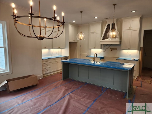 kitchen featuring custom exhaust hood, hanging light fixtures, ornamental molding, a kitchen island with sink, and white cabinetry