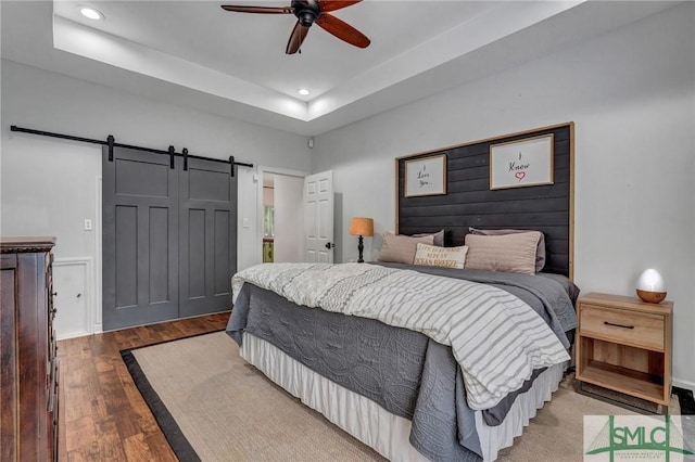 bedroom with a tray ceiling, wood finished floors, recessed lighting, a barn door, and ceiling fan