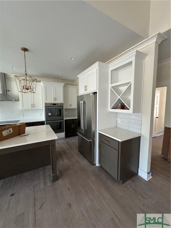 kitchen with high end fridge, hanging light fixtures, double oven, white cabinets, and wall chimney range hood