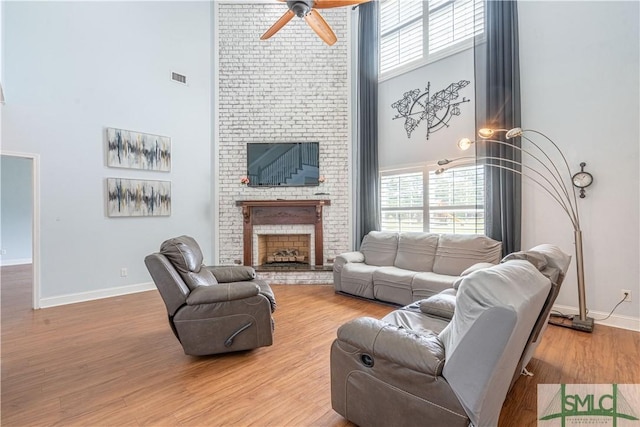 living room with ceiling fan, a fireplace, a towering ceiling, and light hardwood / wood-style floors