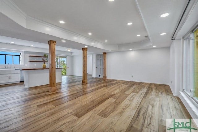 unfurnished living room featuring sink, a raised ceiling, decorative columns, light wood-type flooring, and ornamental molding