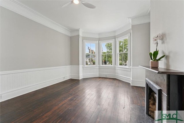 unfurnished living room with ceiling fan, wood-type flooring, a wainscoted wall, crown molding, and a fireplace