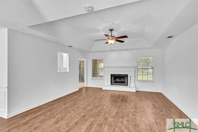 unfurnished living room with a brick fireplace, ceiling fan, wood-type flooring, and a tray ceiling