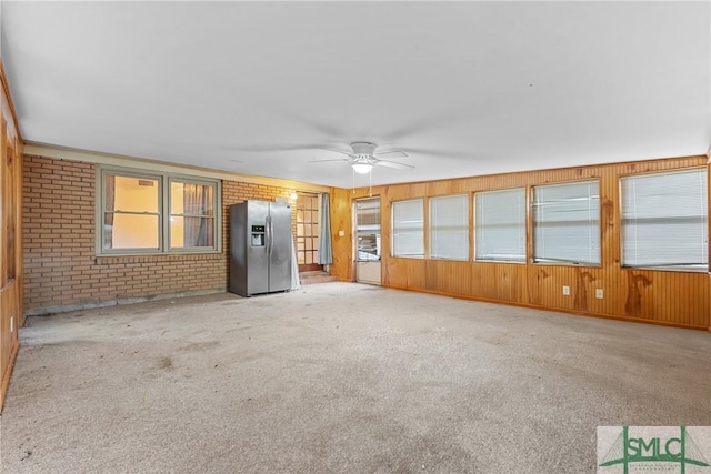 unfurnished living room with wood walls, plenty of natural light, ceiling fan, and light colored carpet