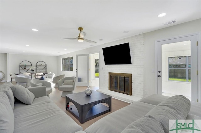 living room featuring ceiling fan, plenty of natural light, wood-type flooring, and a brick fireplace