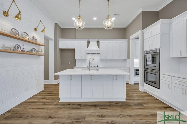 kitchen with white cabinets, a center island with sink, premium range hood, and double oven