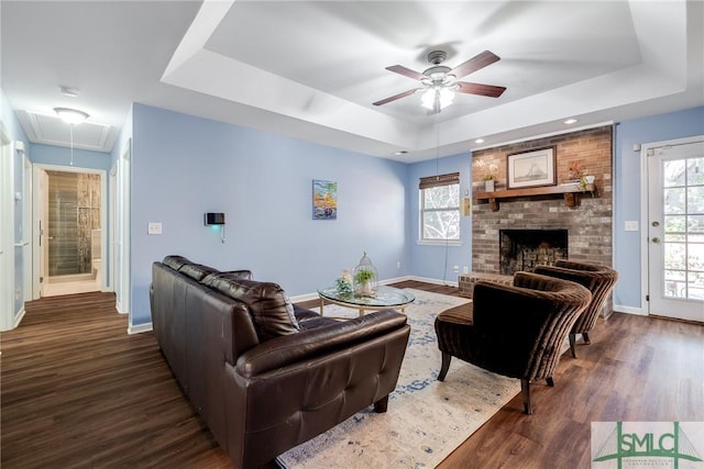 living room with a raised ceiling, ceiling fan, dark hardwood / wood-style floors, and a brick fireplace