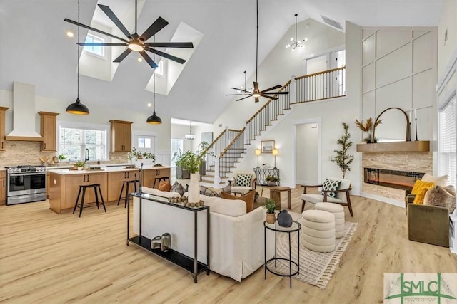 living room with a stone fireplace, a skylight, high vaulted ceiling, light hardwood / wood-style flooring, and ceiling fan with notable chandelier