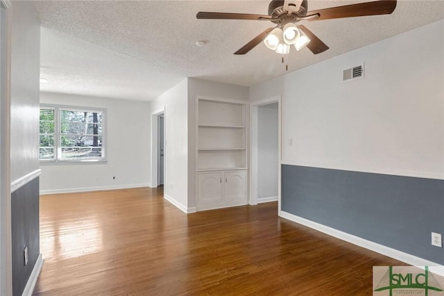 empty room with built in shelves, ceiling fan, a textured ceiling, and dark wood-type flooring