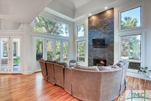 living room featuring french doors, a healthy amount of sunlight, a stone fireplace, a towering ceiling, and light wood-type flooring