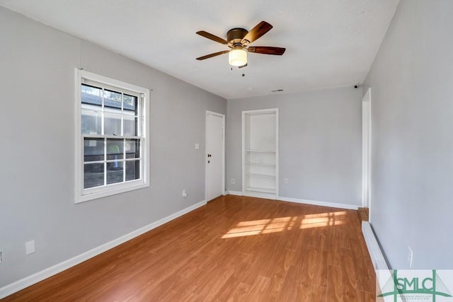 empty room featuring built in shelves, visible vents, a ceiling fan, wood finished floors, and baseboards