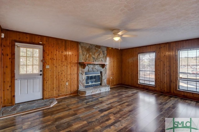 unfurnished living room with dark hardwood / wood-style flooring, a textured ceiling, ceiling fan, a stone fireplace, and wood walls