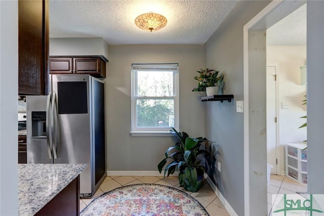 kitchen with stainless steel refrigerator with ice dispenser, light tile patterned floors, a textured ceiling, dark brown cabinets, and light stone counters