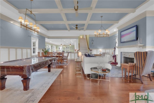 rec room with wood-type flooring, coffered ceiling, beam ceiling, a premium fireplace, and ceiling fan with notable chandelier