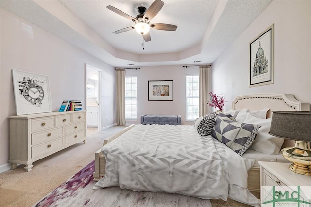 bedroom featuring a textured ceiling, a raised ceiling, ceiling fan, ensuite bath, and light colored carpet