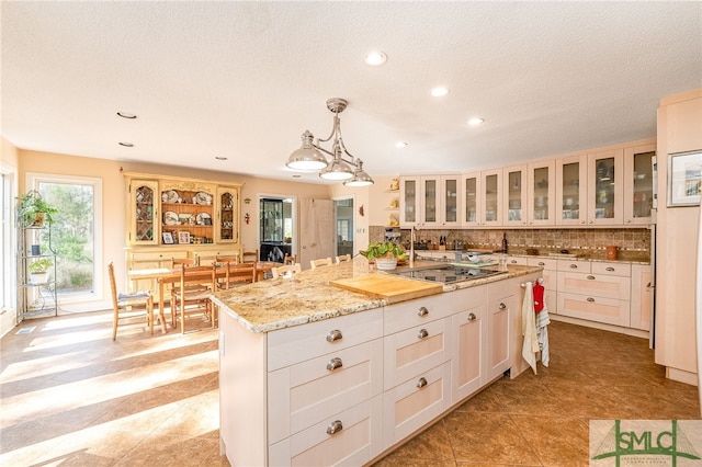 kitchen with hanging light fixtures, black electric stovetop, an island with sink, light stone counters, and tasteful backsplash