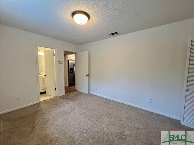 unfurnished bedroom with ensuite bath, a textured ceiling, stainless steel fridge with ice dispenser, and light colored carpet