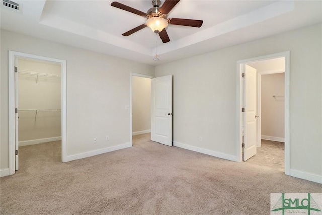 unfurnished bedroom with ceiling fan, a closet, a walk in closet, a tray ceiling, and light colored carpet