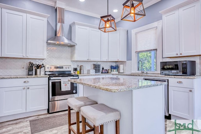 kitchen featuring appliances with stainless steel finishes, a kitchen bar, wall chimney exhaust hood, white cabinetry, and decorative light fixtures