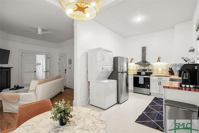 kitchen with stainless steel appliances, wall chimney range hood, stacked washer / drying machine, butcher block countertops, and ceiling fan