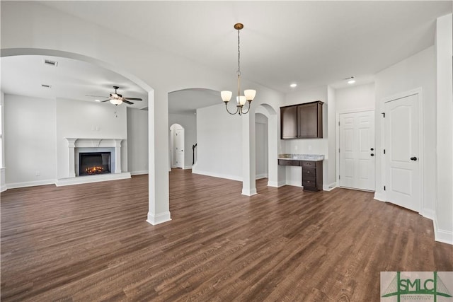 unfurnished living room featuring visible vents, ceiling fan with notable chandelier, a warm lit fireplace, arched walkways, and dark wood-style flooring