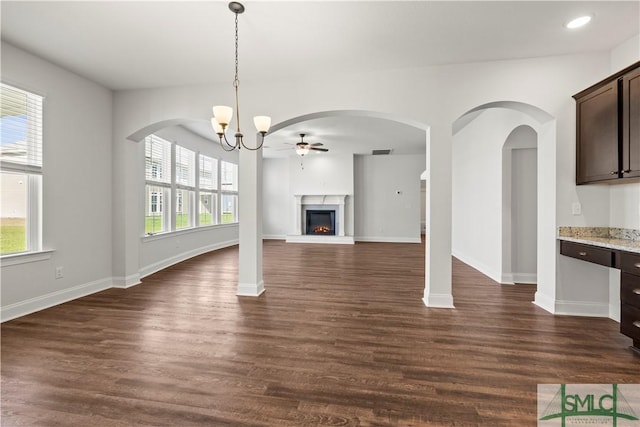 unfurnished living room featuring ceiling fan with notable chandelier, a warm lit fireplace, dark wood-type flooring, and baseboards