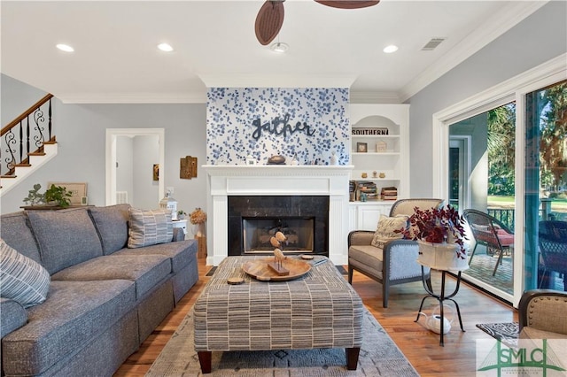 living room featuring a tile fireplace, hardwood / wood-style floors, ceiling fan, ornamental molding, and built in shelves