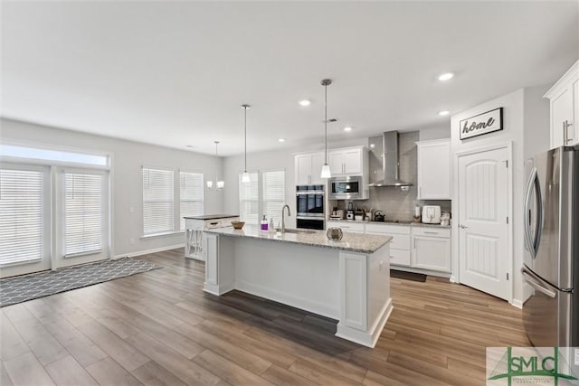 kitchen with white cabinetry, an island with sink, stainless steel appliances, wall chimney range hood, and pendant lighting