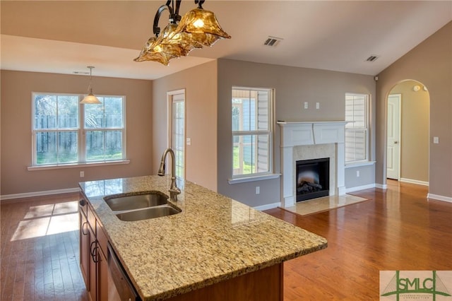 kitchen featuring a fireplace, an island with sink, sink, hanging light fixtures, and light stone counters