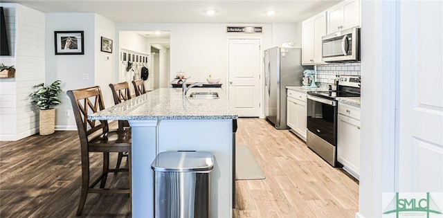 kitchen with white cabinets, appliances with stainless steel finishes, a kitchen bar, a kitchen island with sink, and light stone counters