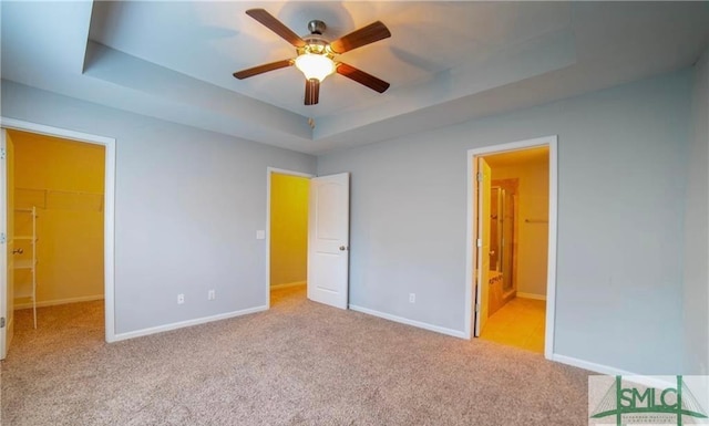 unfurnished bedroom with a walk in closet, light carpet, ceiling fan, connected bathroom, and a tray ceiling