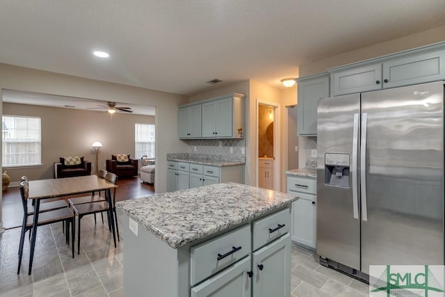 kitchen featuring a kitchen island, stainless steel fridge, backsplash, ceiling fan, and light stone counters