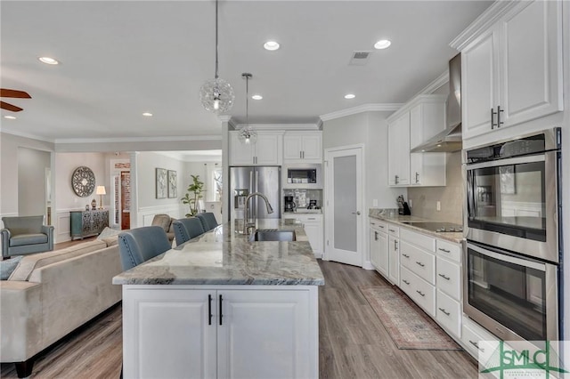 kitchen with appliances with stainless steel finishes, white cabinetry, an island with sink, decorative light fixtures, and wall chimney exhaust hood