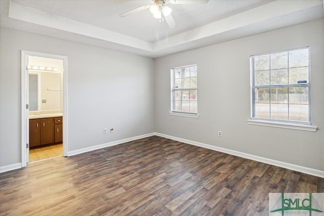 unfurnished bedroom featuring hardwood / wood-style flooring, ceiling fan, ensuite bath, and a tray ceiling