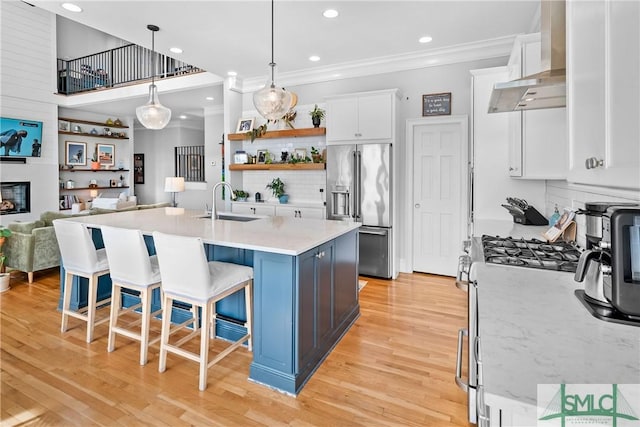 kitchen featuring white cabinetry, hanging light fixtures, an island with sink, stainless steel appliances, and wall chimney range hood