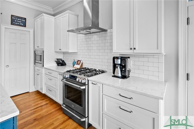 kitchen featuring appliances with stainless steel finishes, white cabinets, ornamental molding, wall chimney range hood, and light hardwood / wood-style flooring