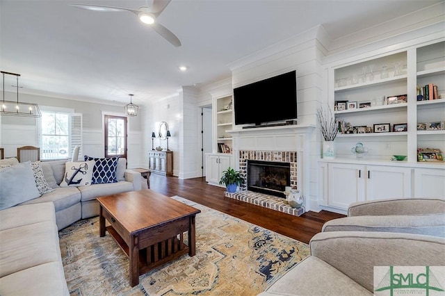 living room featuring built in features, ceiling fan, crown molding, a brick fireplace, and dark wood-type flooring