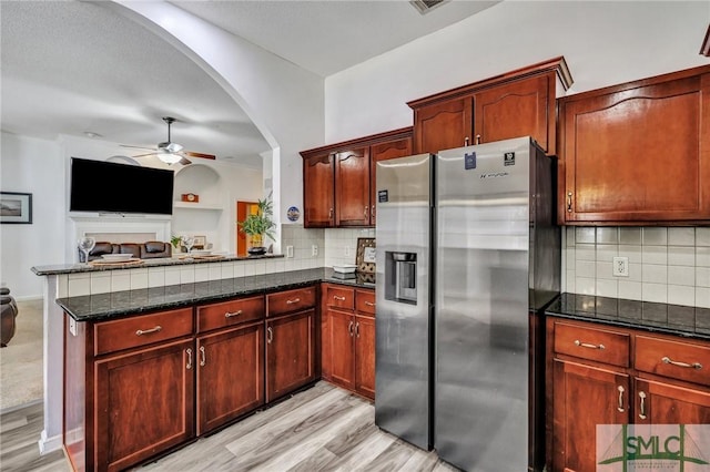 kitchen with stainless steel fridge, ceiling fan, light hardwood / wood-style floors, kitchen peninsula, and dark stone counters