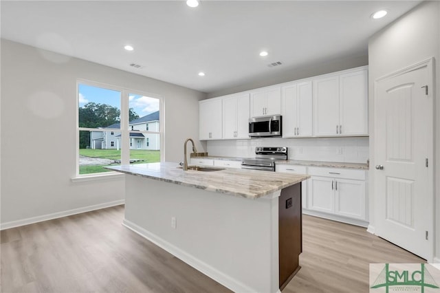 kitchen with sink, appliances with stainless steel finishes, a kitchen island with sink, light stone countertops, and white cabinets