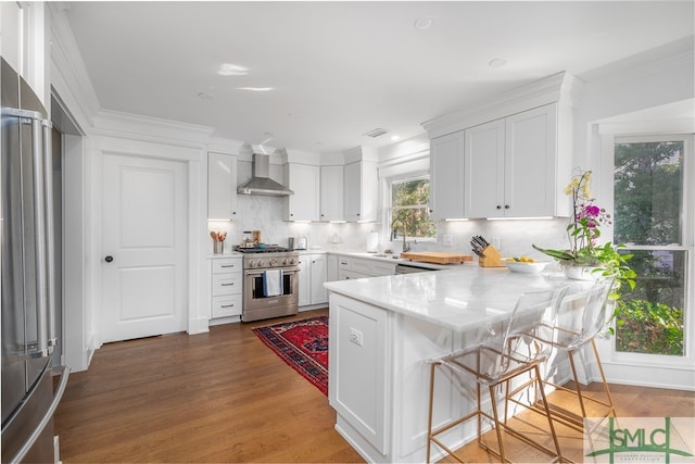 kitchen with a peninsula, appliances with stainless steel finishes, wall chimney range hood, and white cabinets