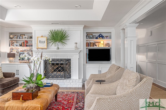 living area featuring built in shelves, a decorative wall, a brick fireplace, a tray ceiling, and ornate columns