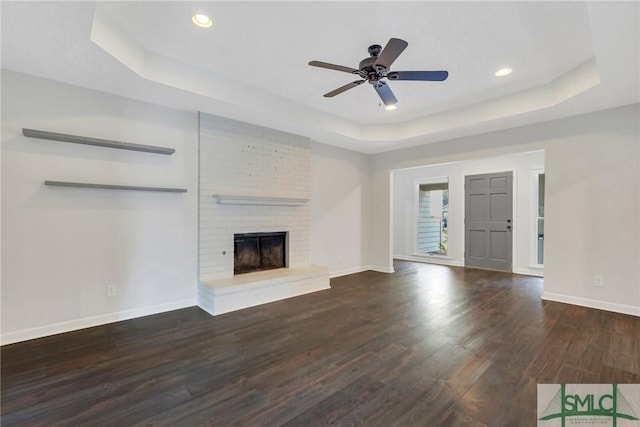 unfurnished living room with a brick fireplace, a tray ceiling, dark hardwood / wood-style floors, and ceiling fan