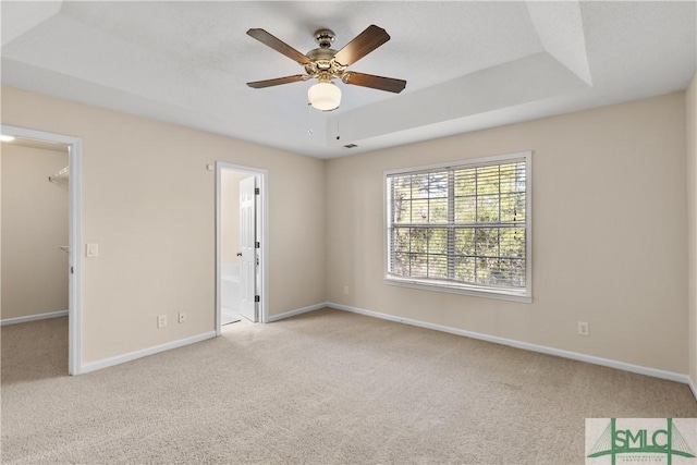 unfurnished bedroom featuring connected bathroom, a spacious closet, light colored carpet, ceiling fan, and a tray ceiling