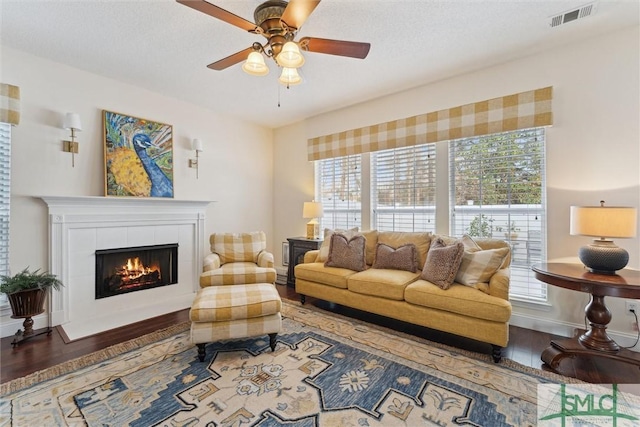 living room with ceiling fan, wood-type flooring, a tiled fireplace, and a textured ceiling