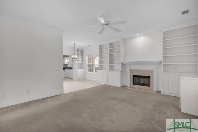 unfurnished living room featuring a tiled fireplace, ceiling fan with notable chandelier, light colored carpet, and built in shelves