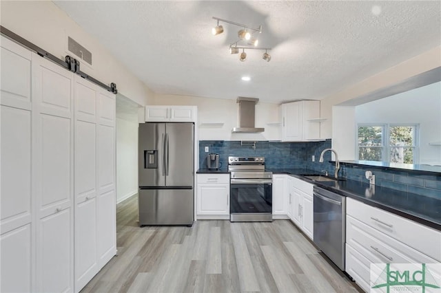 kitchen featuring wall chimney range hood, sink, appliances with stainless steel finishes, white cabinetry, and a barn door