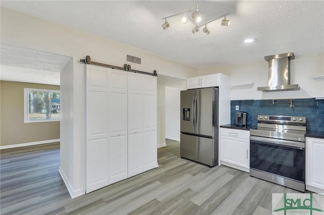 kitchen with wall chimney range hood, stainless steel appliances, white cabinets, a barn door, and light wood-type flooring