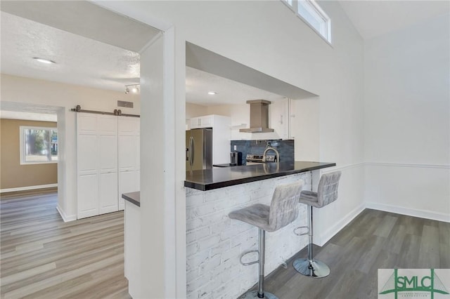 kitchen featuring stainless steel fridge, kitchen peninsula, white cabinets, a barn door, and wall chimney range hood