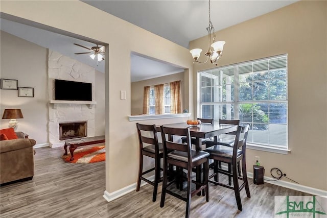 dining area featuring lofted ceiling, a stone fireplace, wood-type flooring, and ceiling fan with notable chandelier