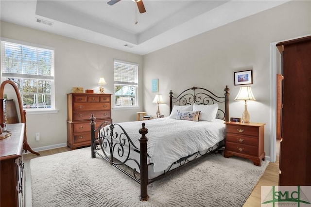 bedroom featuring a raised ceiling, ceiling fan, multiple windows, and light hardwood / wood-style floors